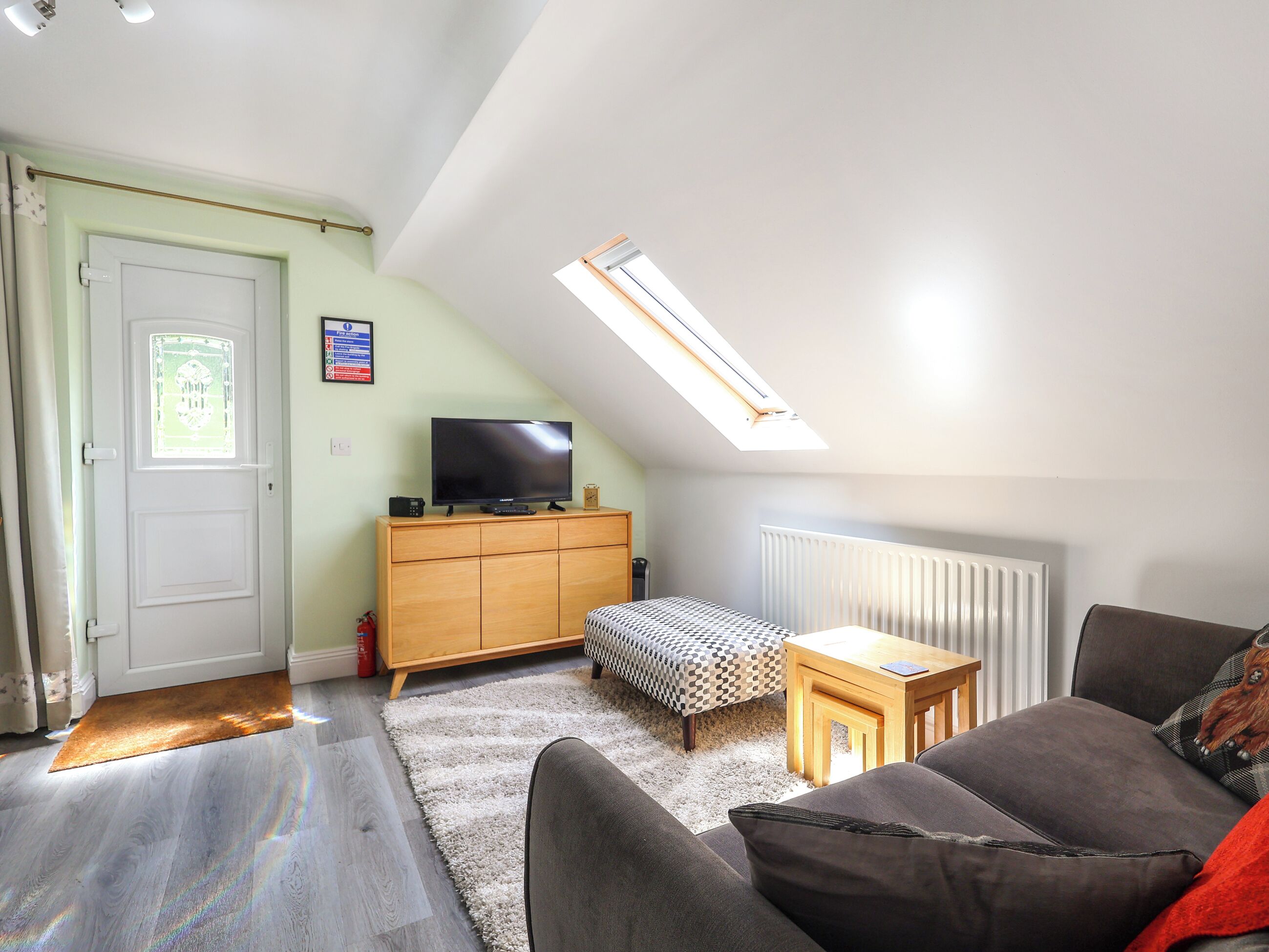 Ett vardagsrum med ett sluttande tak, en tv-apparat placerad på ett träskåp, en mönstrad pall, ett litet träbord och en grå soffa.