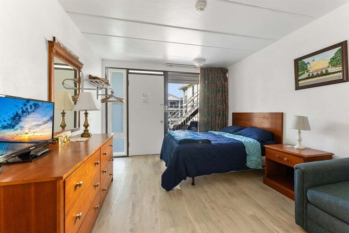A hotel room with a bed, a desk with a television, a dresser, and a window with a view of a building.