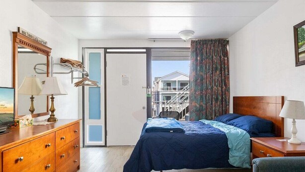 A hotel room with a bed, dresser, mirror, and a view of a building outside. - Wildwood Studio Condo with a Pool (Wildwood)
