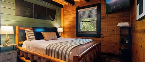 A wooden cabin bedroom with a bed, nightstands, a window with blinds, and a television on the wall.