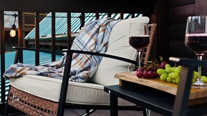 Una silla de mimbre con un cojín y una manta, una mesa con una copa de vino tinto y uvas, y una vista de un edificio con tejado azul.