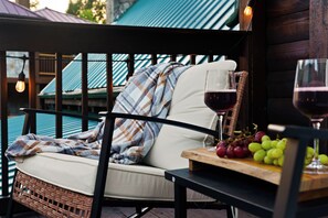 A wicker chair with a cushion and a blanket, a table with a glass of red wine and grapes, and a view of a building with a blue roof.