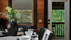 A dining area with a table set for two, a vase with flowers, and a view of greenery through a door.