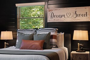 A bed with multiple pillows, a window with blinds, and a wall sign that reads 'Dream & Sweet'.