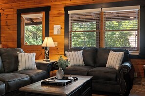 Ein gemütliches Wohnzimmer mit einer Ledersofa, einem Couchtisch und einer Lampe. Zwei Fenster mit Jalousien, und ein Zettel an der Wand.