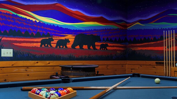 Una mesa de billar con un mural colorido de osos y un cielo nocturno, bancos de madera y un soporte para tacos.