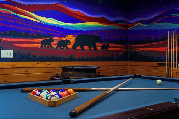 A pool table with a colorful mural of bears and a night sky, wooden benches, and a pool cue rack.