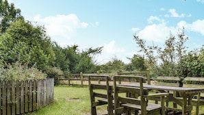 A wooden picnic table and benches in a grassy area with a wooden fence and trees in the background. - 2 bedroom accommodation in Camelford (Camelford)