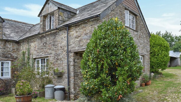 A stone building with a sloped roof, a large bush in front, and a wheelbarrow to the left. - 2 bedroom accommodation in Camelford (Camelford)