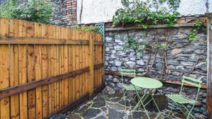 A stone patio with a wooden fence, a green metal table and chairs, and a stone wall with climbing plants. - 1 bedroom accommodation in Keswick (Keswick)