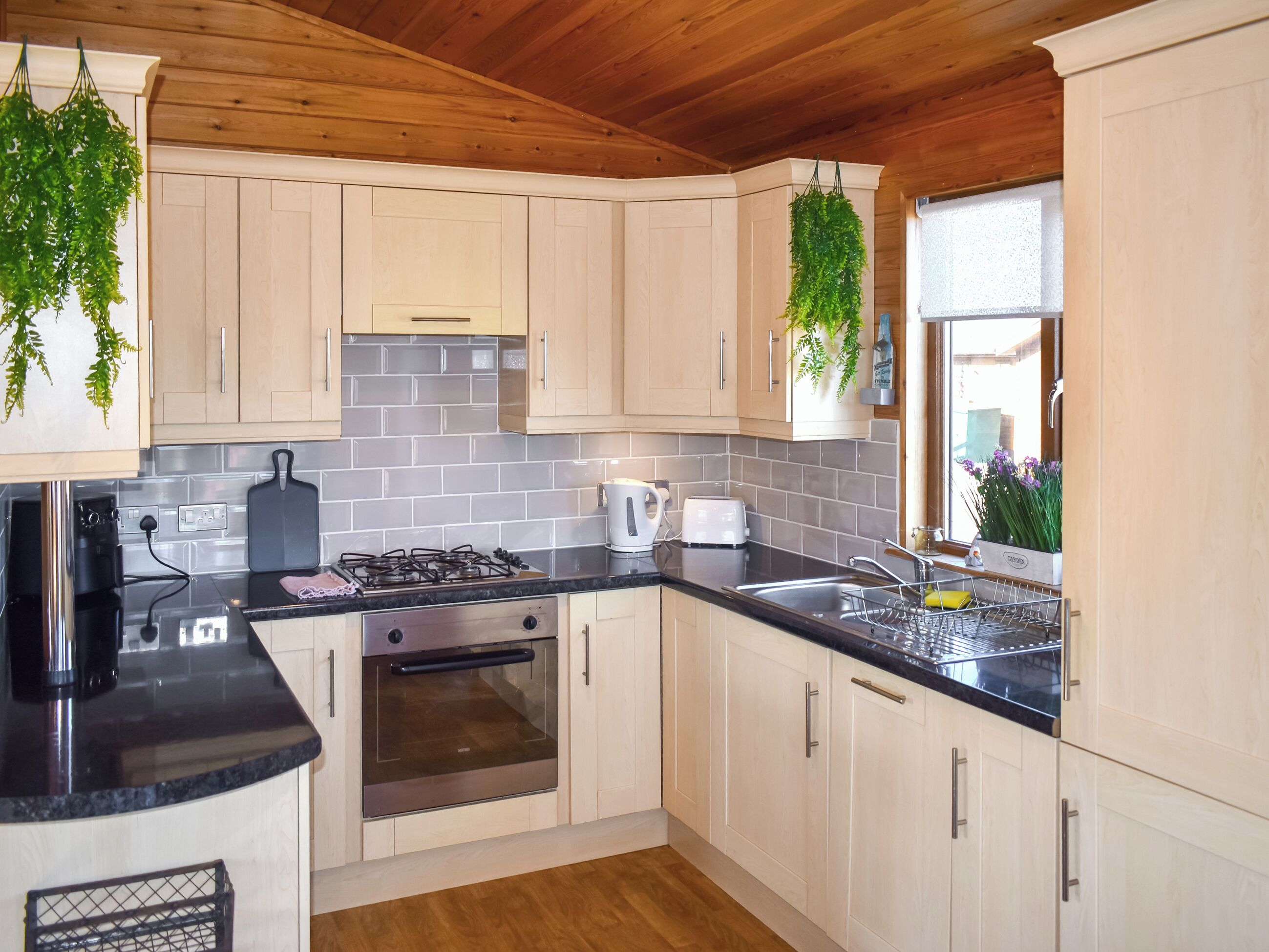 A modern kitchen with light wooden cabinets, a black countertop, a gas stove, an oven, and hanging plants.