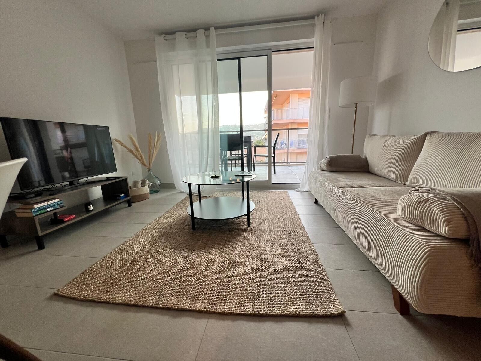 Een moderne woonkamer met een flatscreen televisie, een glazen salontafel, een beige bank en een groot raam met uitzicht op een balkon.