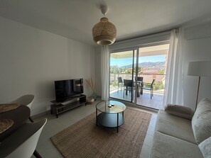 En moderne stue med et fladskærms-tv, et rundt sofabord og en sofa. Der er en balkon med udsigt over byen og bakker.