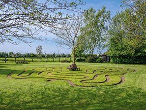 Ein grünes Labyrinth mit einem Baum in der Mitte, Picknicktischen und einem Holzzaun im Hintergrund.