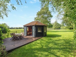 A wooden gazebo with a tiled roof, a wooden deck with chairs, and a grassy field with a fence in the background.