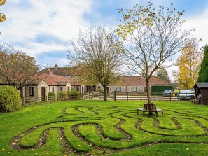 A stone building with a tiled roof, surrounded by a grassy maze, a wooden bench, and a tree with yellow leaves.