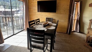 A dining area with a wooden wall, a flat-screen TV mounted above a table with four chairs, and a large window with a view of trees and buildings.