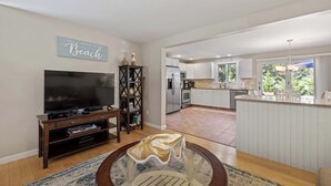 Un salón con televisor, centro de entretenimiento y comedor con una mesa redonda. Se ve una cocina con armarios blancos y un refrigerador al fondo.