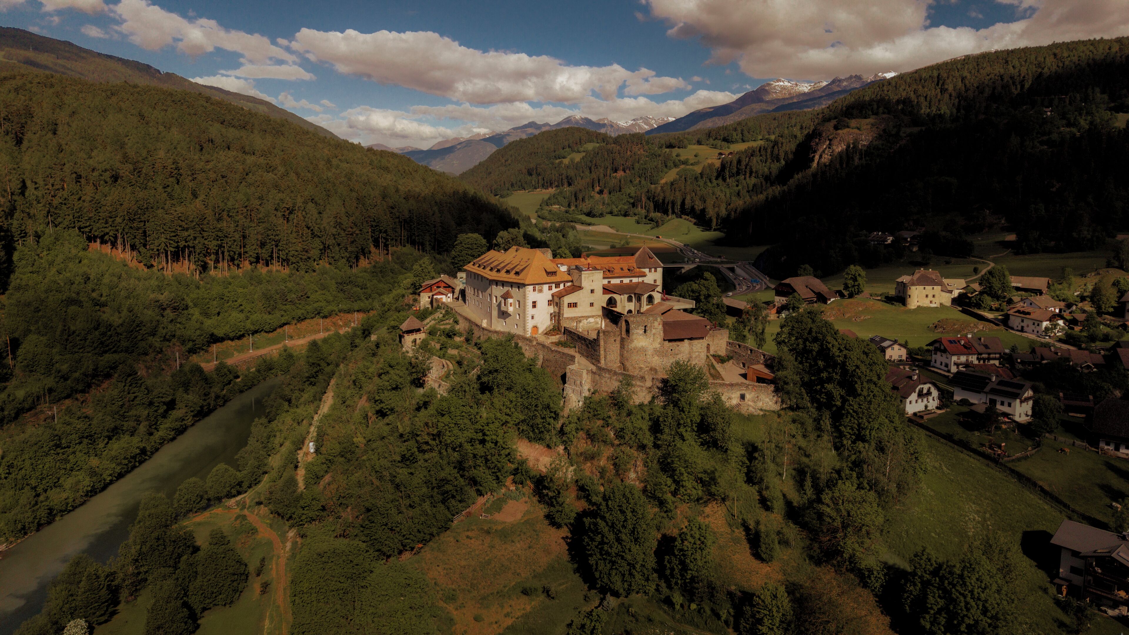 A castle-like structure with a river, surrounded by forested hills and mountains.
