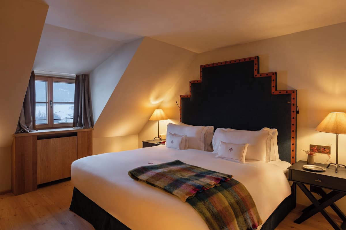 A hotel room with a large bed, a wooden desk, two bedside lamps, and a window with curtains.