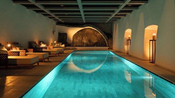 A modern indoor pool with a clear blue water feature, a stone wall, and seating areas with soft lighting. - Castel Badia (San Lorenzo di Sebato)