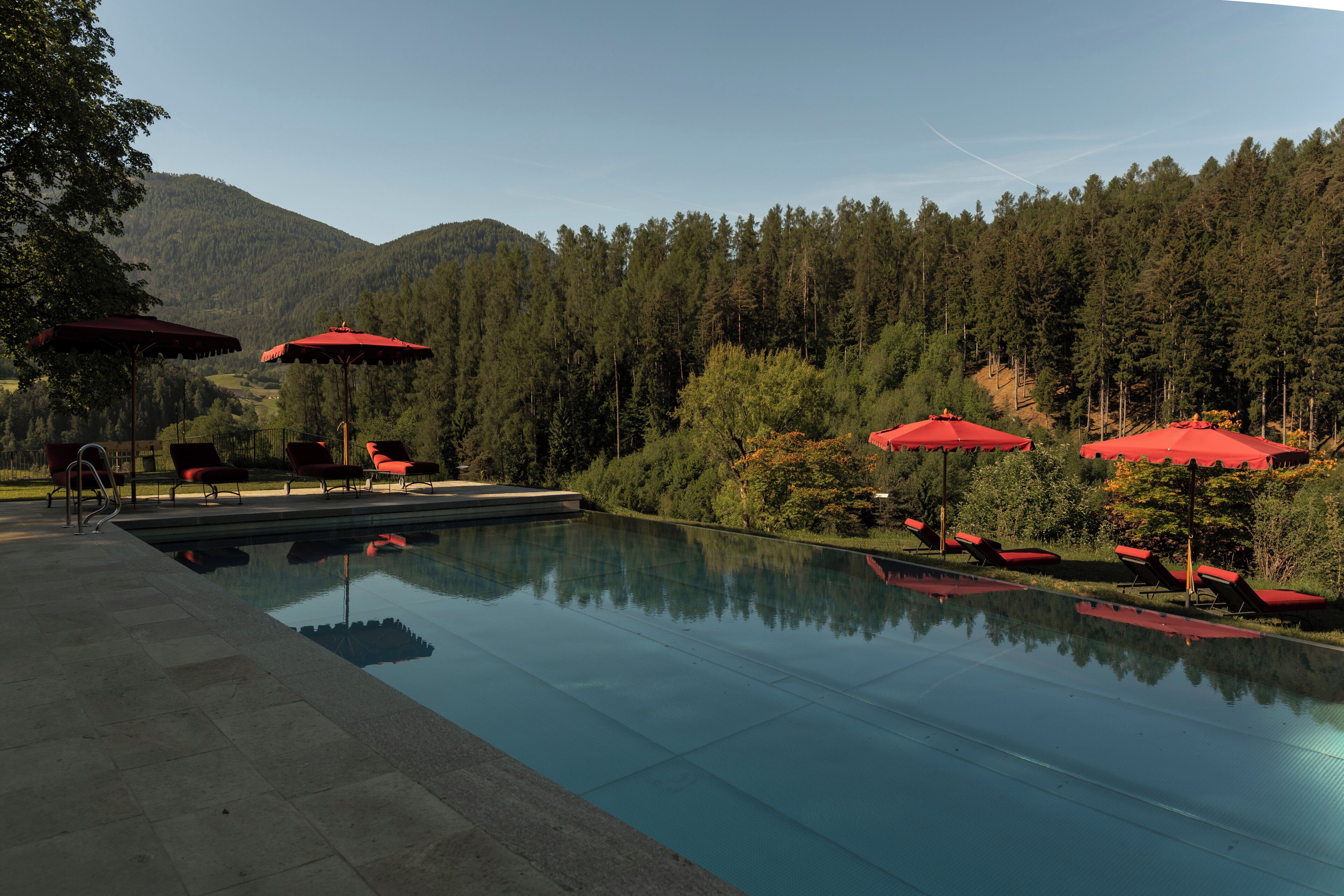 A swimming pool with red umbrellas and lounge chairs, surrounded by a forested hillside.