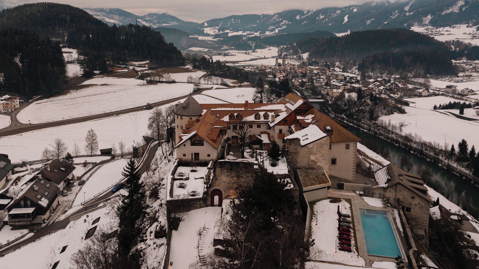 A historic building with a courtyard and a swimming pool, surrounded by snow-covered landscapes and mountains.