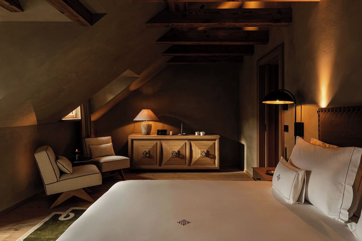 A cozy, dimly lit bedroom with a large bed, a small desk, a chair, and a lamp.