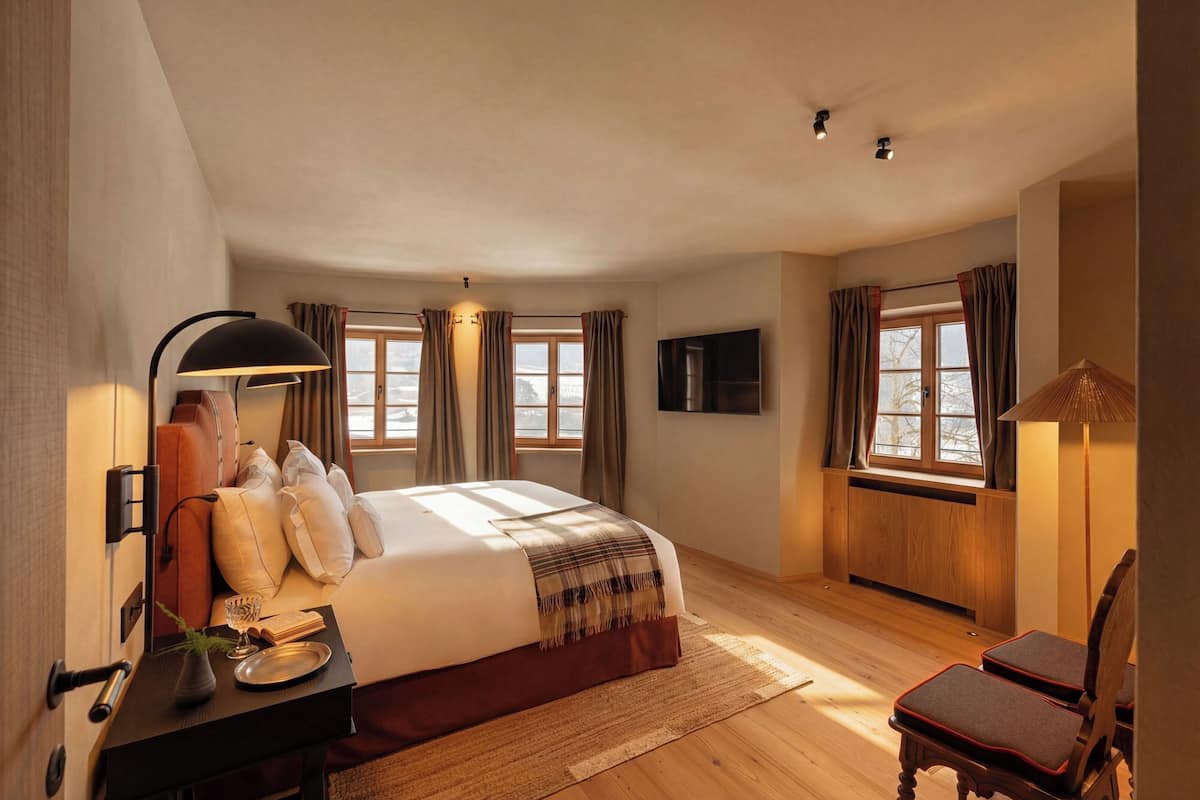 A hotel room with a large bed, a bedside table with a lamp, a TV mounted on the wall, and a window with curtains.