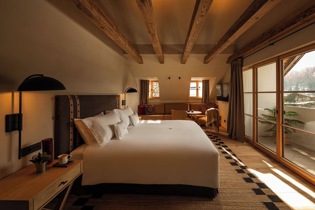 A large bed with a headboard, a bedside table with a lamp, a small plant, and a cup. There is a window with curtains and a view of the outside.