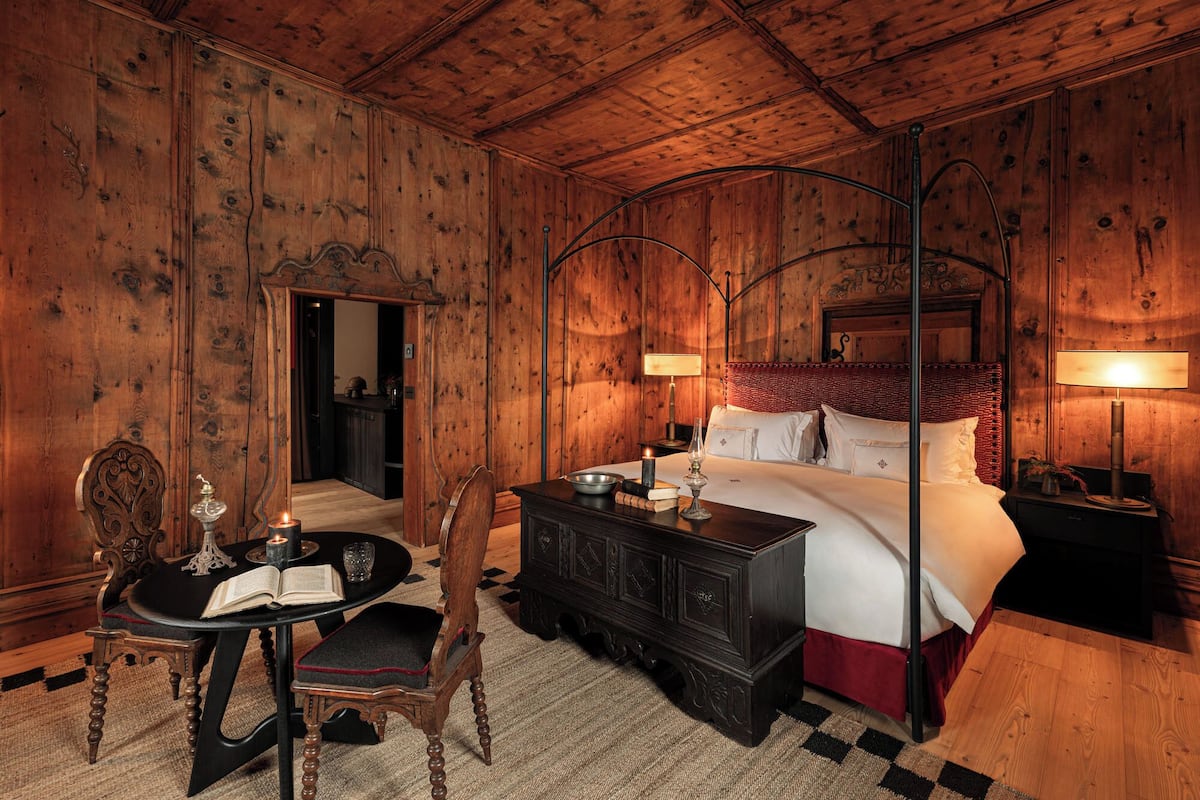 A wooden-paneled bedroom with a four-poster bed, a table with a book and candle, and a chair.