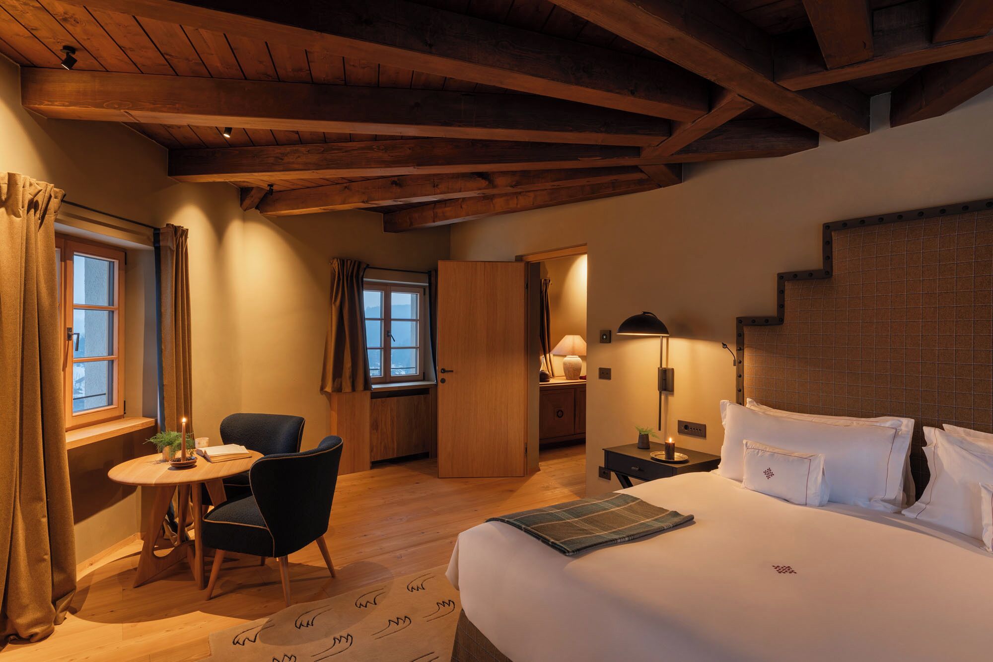 A cozy hotel room with a wooden ceiling, a bed with white linens, a small round table with a chair, and a window with curtains.