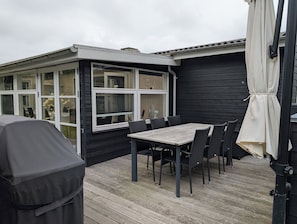 A covered outdoor patio with a table and chairs, a grill, and a view of a building with large windows. - Charming cottage with large fenced garden (Løgstør)