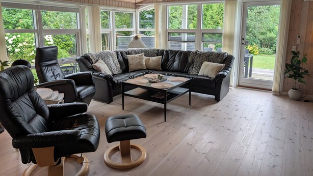 A modern living room with a leather sofa, black armchairs, a coffee table, and a large window offering a view of greenery. - Charming cottage with large fenced garden (Løgstør)