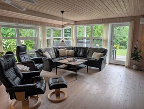 A spacious living room with a leather sofa, a coffee table, and a black armchair. Large windows provide a view of the outdoors. - Charming cottage with large fenced garden (Løgstør)