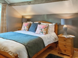 A bedroom with a wooden bed, floral pillows, a blue blanket, a wooden nightstand with a lamp, and a small drawer.