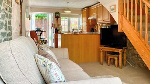 A living room with a sofa, a television on a wooden stand, and a kitchen area with wooden cabinets and a window. - The Old Post House Annex - UK50190 (Axbridge, near Weare)