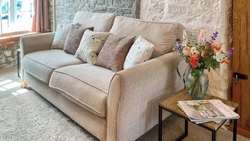 Ein beiges Sofa mit gemusterten Kissen, ein Couchtisch aus Holz mit einer Zeitung und eine Vase mit Blumen.