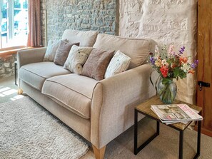 Un canapé beige agrémenté de coussins à motifs, une table basse en bois sur laquelle repose un journal et un vase rempli de fleurs.