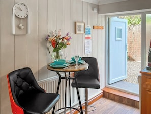 A small dining area with two black chairs, a round table, and a vase of flowers. There is a clock on the wall and a door leading outside.