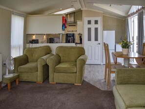 Een compacte woonruimte met twee groene fauteuils, een bijzettafeltje met een plant in pot, een eethoek met houten stoelen en een kitchenette op de achtergrond.