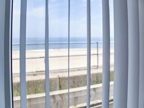 Uitzicht door een raam met verticale jaloezieën, op een strand met zand en een rustige zee.