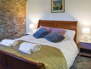 A neatly made bed with a green quilt, two pillows, and rolled towels. There is a wooden headboard, a stone wall, and a painting on the wall.
