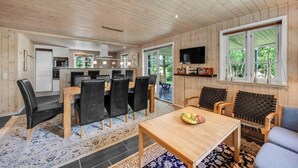 Un vaste espace de vie doté d’un plafond en bois, d’une table à manger avec des chaises, d’un téléviseur fixé au mur et d’une grande fenêtre donnant sur un paysage verdoyant.