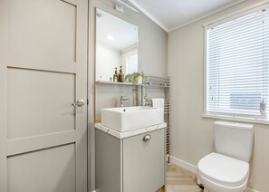 Ein Badezimmer mit einem weißen Waschbecken, einem Spiegel, einer Toilette, einem Handtuchhalter und einem Fenster mit Jalousien.