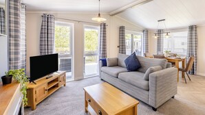 A modern living room with a sofa, TV, wooden table, and a balcony with curtains. - Monument Lakeside Lodge Pet Friendly (Woodhall Spa)