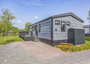 Ein eingeschossiges Mobilheim mit Veranda, einer kleinen Terrasse und einer Auffahrt mit Kies.