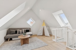 Une chambre à coucher aménagée dans un grenier, avec un plafond en pente, un canapé, une table ronde en bois, une tente blanche et une structure blanche ressemblant à une échelle.