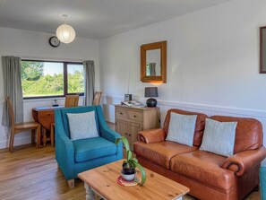 A cozy living room with a blue armchair, a brown leather sofa, a wooden coffee table, and a view of greenery outside. - 2 bedroom accommodation in Isle of Whithorn (Isle of Whithorn)
