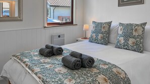 Una cama bien hecha con ropa de cama floral, una mesita de noche con una lámpara, y una ventana con vista a un edificio.